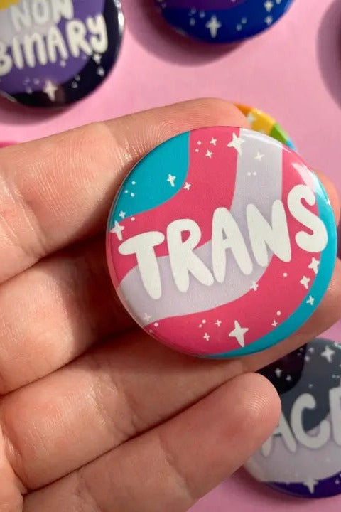 Hand holding the round Trans pride flag identity button in pink, blue and white with sparkles and hand-lettered text
