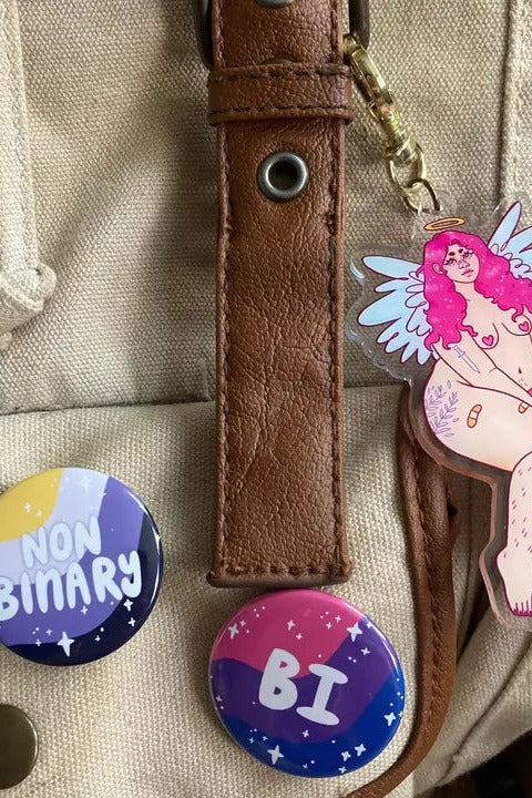 Nonbinary and Bi pride flag identity buttons pinned to a canvas backpack strap, styled with an illustrated acrylic charm