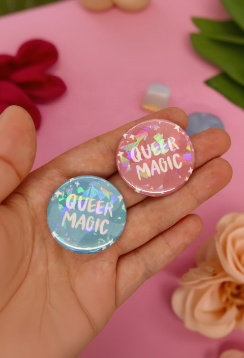 Hand holding two Queer Magic holographic pinback buttons in pastel pink and blue with white lettering, with flowers and crystals on pink