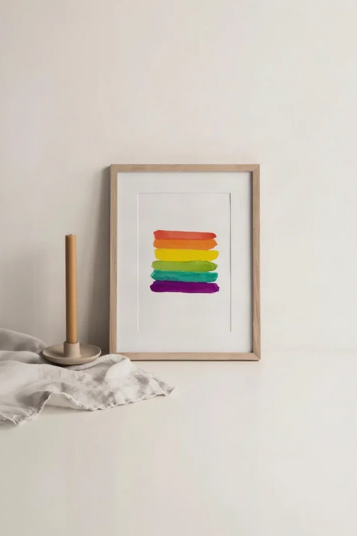 Watercolor print of six stacked rainbow brush strokes in red, orange, yellow, green, teal and purple, in a light wood frame leaning against a cream wall