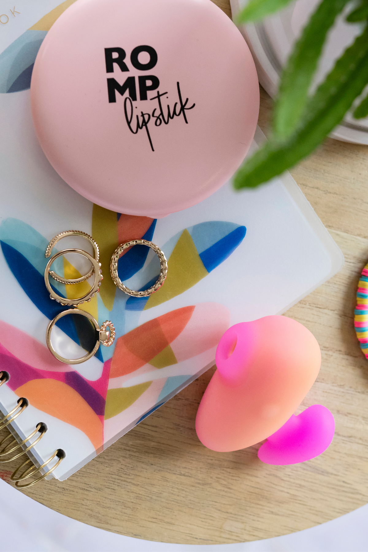 Romp Kiss suction toy lying on a dresser beside a pink Romp lipstick compact, rings and a leafy plant, a lifestyle shot of the discreet travel vibe