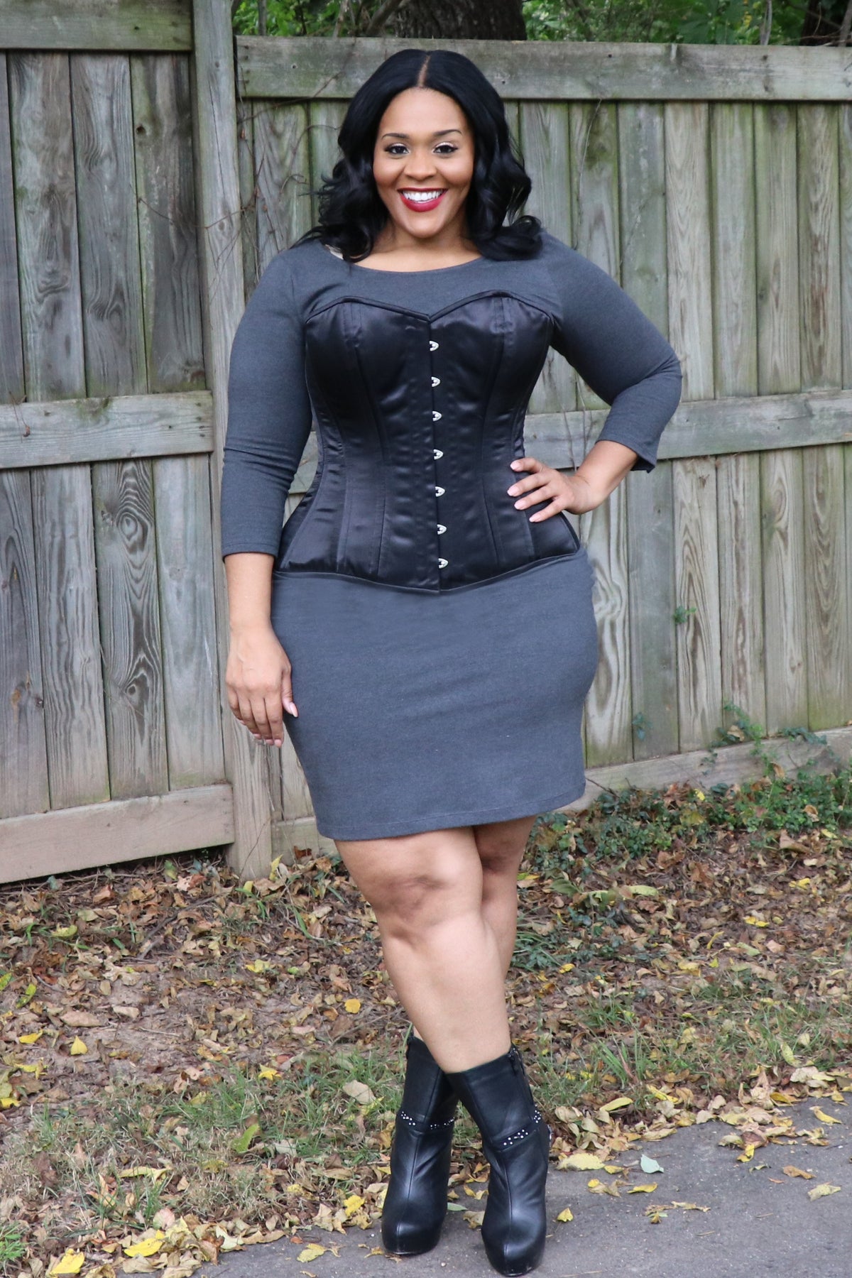 Model in a grey dress and black boots wearing the black Seductress overbust corset cinched at the waist, posed outdoors by a wooden fence