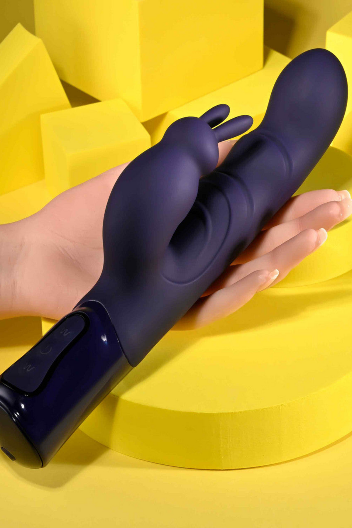Hand holding the purple She's A Big Deal Rabbit Vibe against yellow studio blocks, showing the girthy shaft and rabbit ears next to a palm for scale