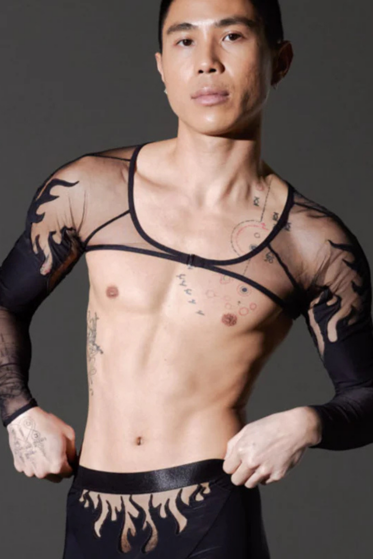 Model wearing the Smokin Mirrors shrug in black, a sheer long-sleeve harness-style top with flame print sleeves, with flame boxer briefs