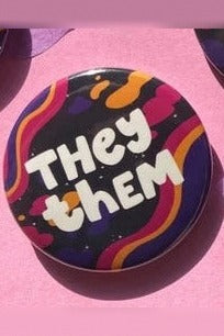 Spacey They/Them pronoun button with white bubble lettering over orange, pink and purple wavy stripes on a dark background, on pink felt