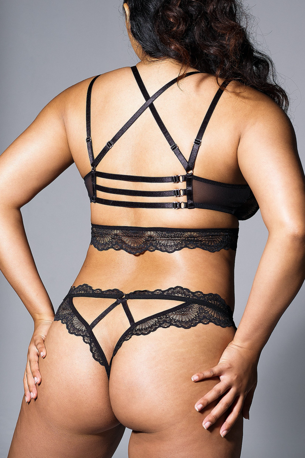Back view of a model in the Sugar and Kane black crotchless lace bikini with strappy cutout back, worn with a matching strappy black bra