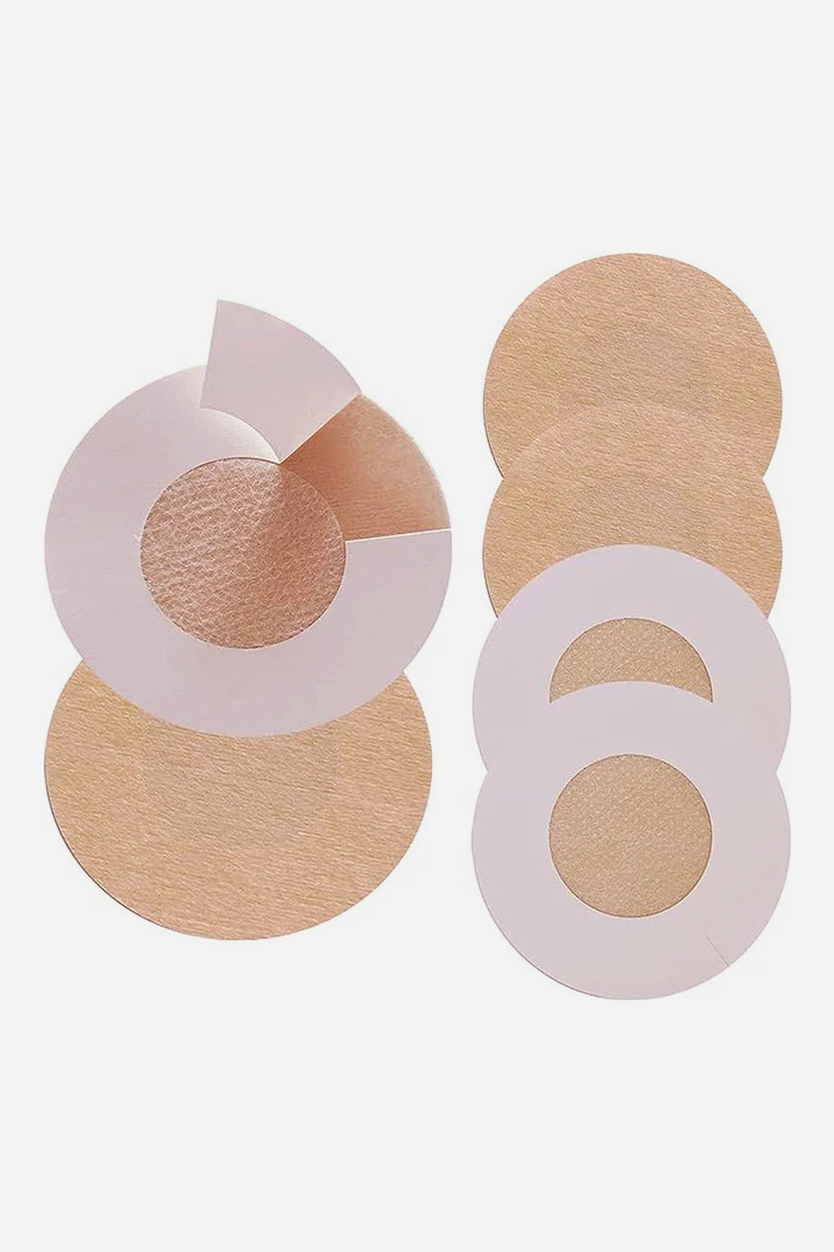 Dark beige round fabric nipple covers shown with their white backing liners, one cover partly peeled off its liner to reveal the adhesive