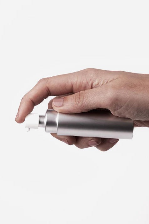 Hand holding the silver Uberlube Good to Go case sideways with the spray nozzle pointing left, showing its slim travel size on light grey