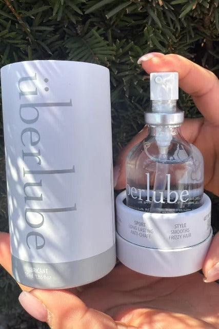 Hand holding an Überlube silicone lube bottle in its white cylindrical tube box against green foliage in sunlight