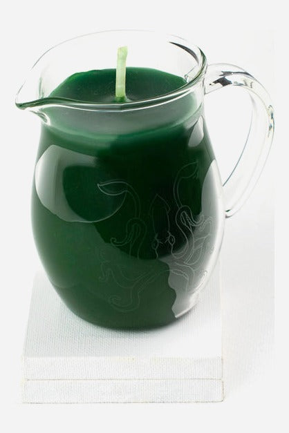 Green wax play pitcher candle in a clear glass jug with handle and pour spout, set on a small white block against light grey