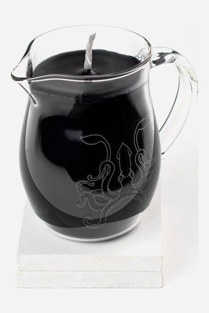 Black wax play pitcher candle in a clear etched glass jug with handle and pour spout, set on a small white block against light grey