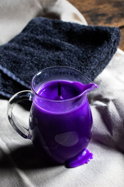 Purple wax play pitcher candle with a drip of melted wax, resting on a white sheet beside a folded dark towel on a wooden surface