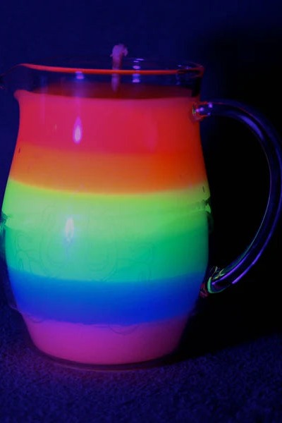 Close view of the glass pitcher candle under UV light with bands of fluorescent orange, yellow, green, blue and pink wax glowing in the dark