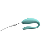 hands-free wearable c-shaped aqua We-Vibe sync lite with the pin charging cord close to the toy, showing the toy is rechargeable. Pin chargers often mean the toy is not waterproof, as in this case