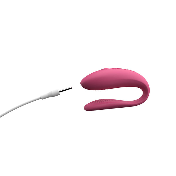 hands-free wearable c-shaped pink We-Vibe sync lite with the pin charging cord close to the toy, showing the toy is rechargeable. Pin chargers often mean the toy is not waterproof, as in this case