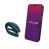 c-shaped wearable sex toy in green velvet next to a cell phone with a we-vibe logo on its screen. meant to show that the sex toy can be controlled via the phone app