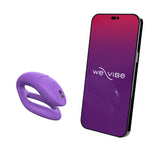 c-shaped wearable sex toy in lilac next to a cell phone with a we-vibe logo on its screen. meant to show that the sex toy can be controlled via the phone app