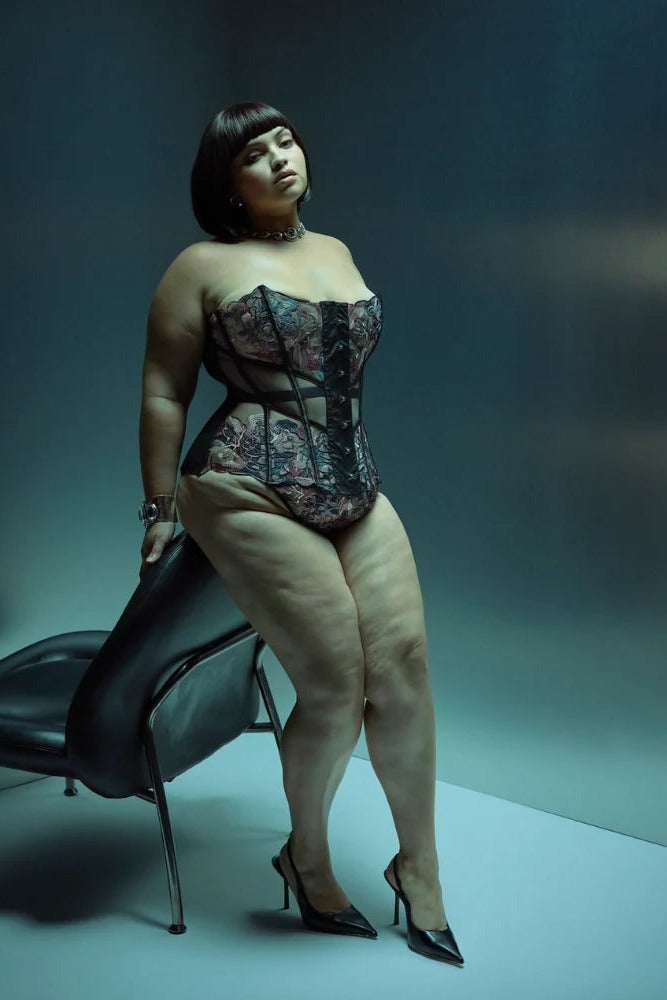 Curvy model in the Wild Chanterelle Dreams corset and heels standing beside a black chair in a teal-lit studio, showing the full-length fit of the corset