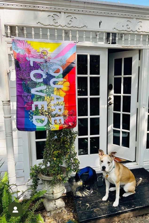 You Are Loved progressive pride flag hanging over the French doors of a white house porch with a dog sitting on the doormat