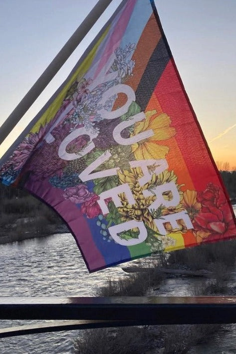 You Are Loved progressive pride flag waving from a pole on a boat at sunset, showing the floral print and white lettering
