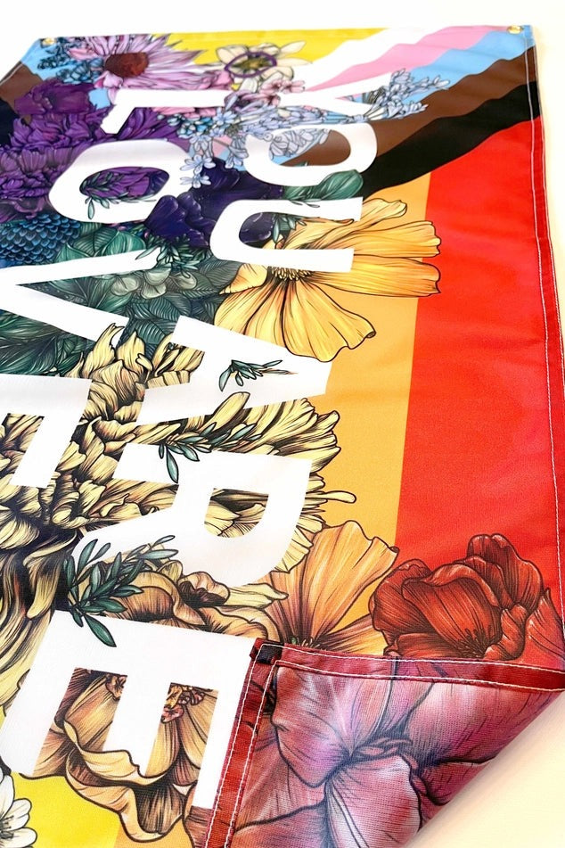 Close-up of the You Are Loved flag's bold white lettering over rainbow stripes and detailed hand-drawn flowers by Transpainter