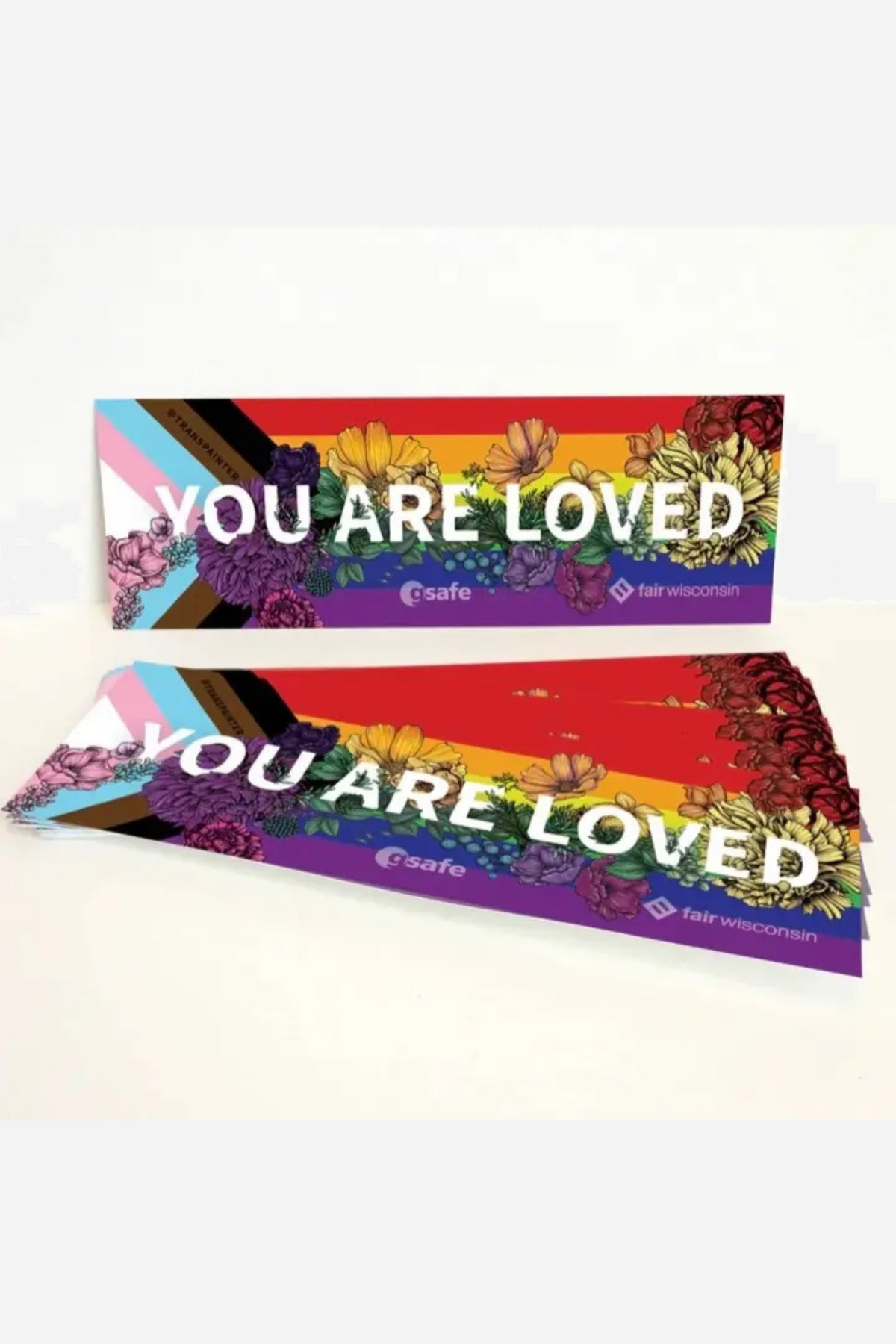 Two You Are Loved bumper stickers stacked at an angle, the words in white over a progress Pride flag scattered with painted flowers.