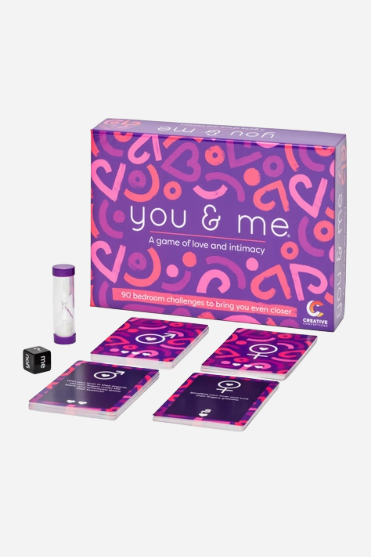 You and Me game box in purple and pink, shown with a sand timer, a die and a few of its challenge cards laid out in front of it.