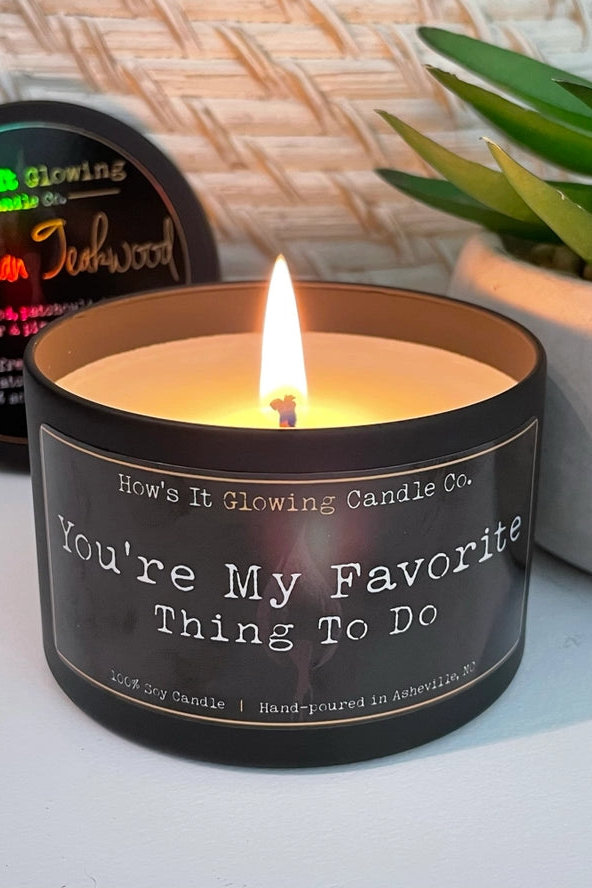 Lit black tin soy candle labelled You're My Favorite Thing To Do by How's It Glowing Candle Co., beside a succulent and another candle tin