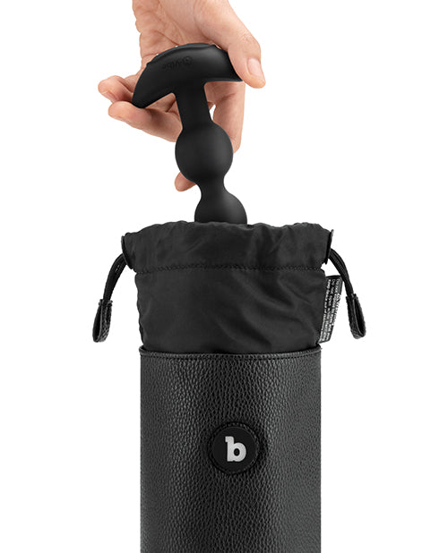 A white person's hand is shown putting anal beads into the black UV sanitizing pouch. The pouch shows a drawstring closure and a button with a lowercase b in the middle of it.