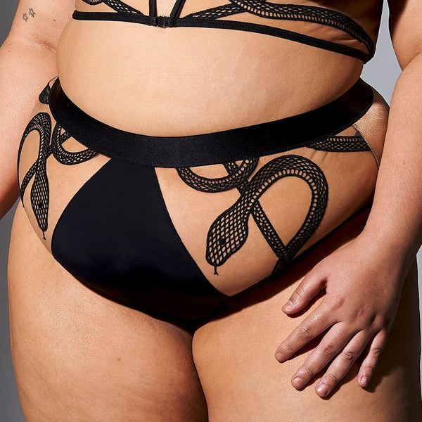 close up of white model wearing high waisted bikini in skintoned mesh with embroidery snakes detail. plus size model