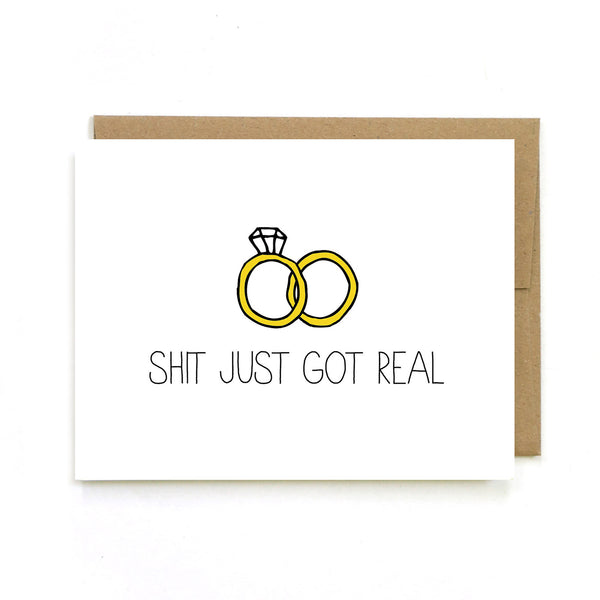 Sex Positive Greeting Card with handwriting style font. A white card with “Shit just got real!” written in all caps black font, above the writing is a diamond ring and a wedding band, slightly overlapping. This card is made by Unblushing a Women-Owned small sex positive graphic designer.